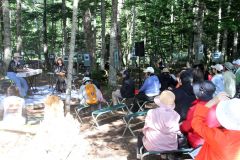 湖のほとりの静かな森林の中で、ゆりずむの演奏に耳を傾ける来場者たち。奥には森の美術館と称し、木々に写真を展示している