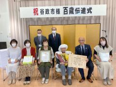 金屏風の前で記念撮影する谷さん（右から３番目）と飯田町長（右から２番目）、後列左から成田総合施設長、野坂理事長