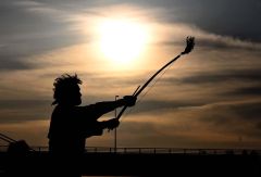 帯広カムイトウウポポ保存会による弓の舞「ク　リムセ」。弓を掲げる男性を夕日が照らす。狩人が鳥を撃とうとした時、鳥が美しく撃つことができなかったという物語からこの踊りができたと伝わる。神々への奉納の踊りとして儀式や祭事の時に踊られるほか、近年では男性の代表的な踊りとして公演などで披露されている