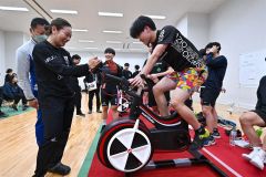 ３０分間で２０キロの距離をこぐワットバイクリレーに参加するスケート部員と見守る（左から）佐藤選手と梅川選手