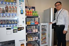 左の自動販売機で決済し購入できる冷凍食品を紹介する泉川事務長