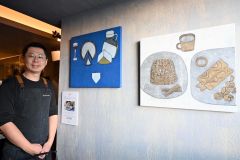 店内の壁に飾った作品の横に立つマリヨンヌの大久保康生オーナー